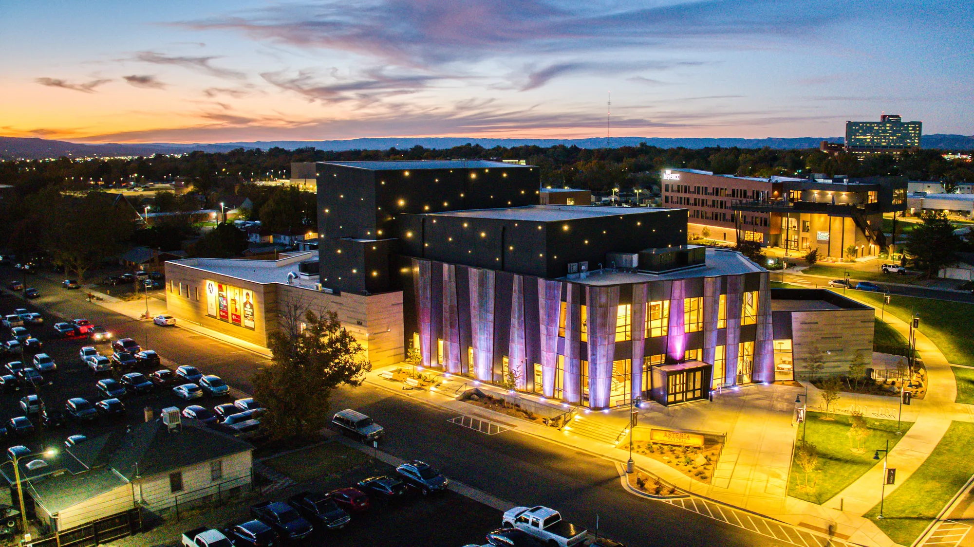 Aerial view at dusk. The lobby glows amber through the glazing and the steel fins are washed in violet light; the dark fly tower behind is dotted with small points of light; the plaza and paths are lit warm. A car park with parked cars fills the foreground, town lights and a long mesa line sit on the horizon under an orange and violet sky. No people visible.